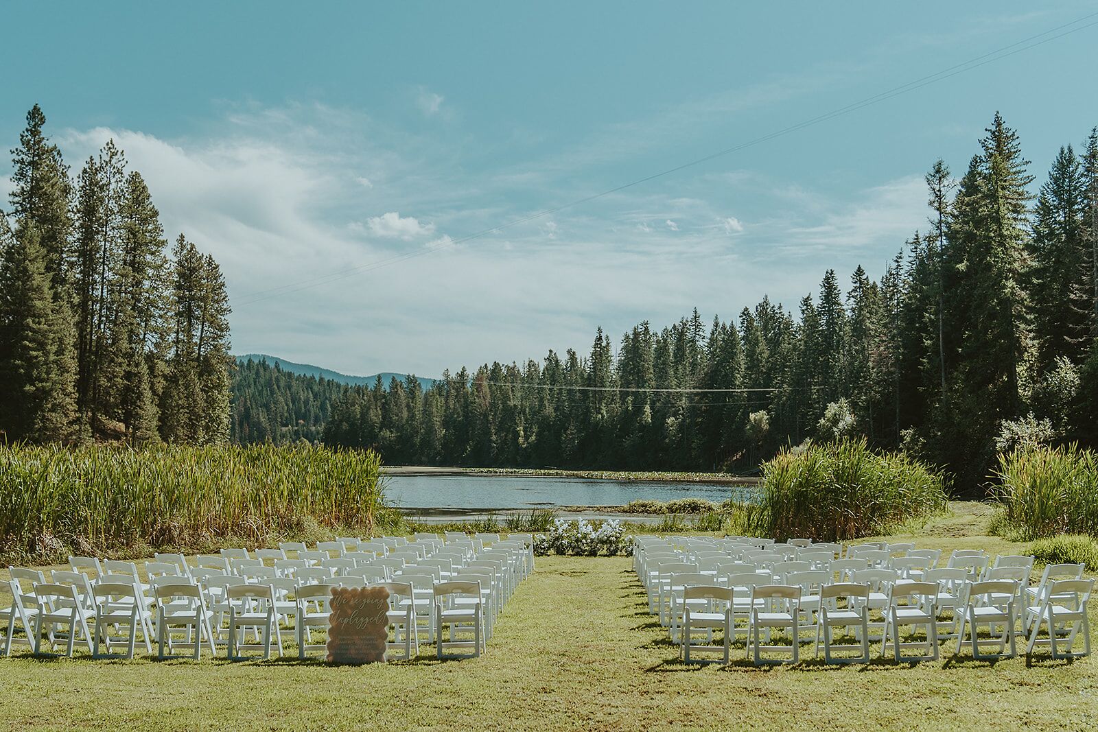 An outdoor wedding venue with rows of white chairs facing a lake, surrounded by dense forest and hills.