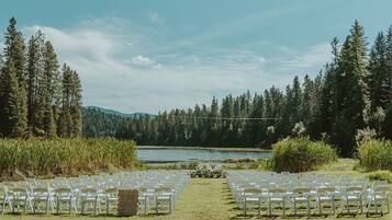 An outdoor wedding venue with rows of white chairs facing a lake, surrounded by dense forest and hills.