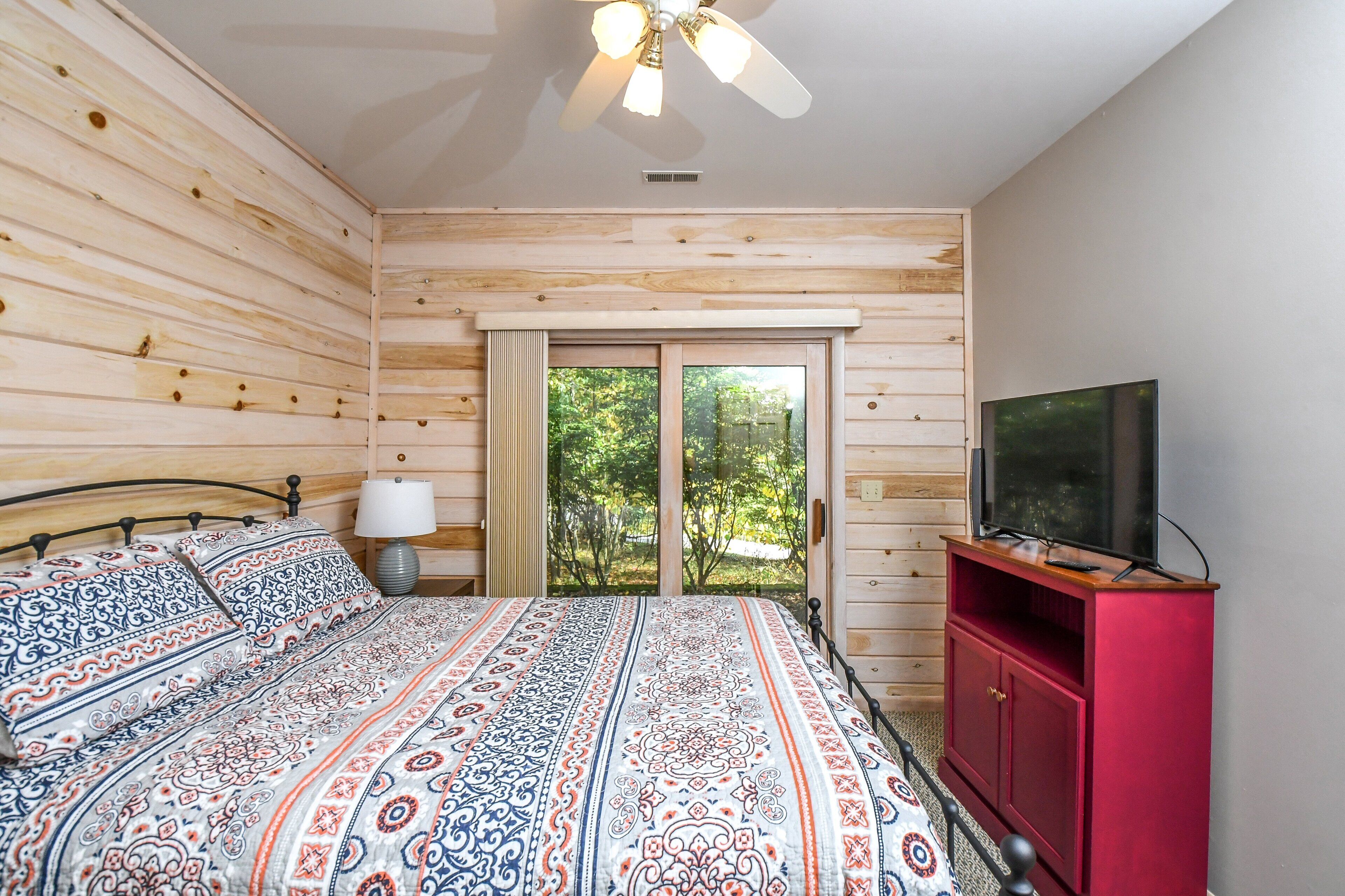A bedroom with a wooden slat wall, a bed with patterned bedding, a red TV stand, and a ceiling fan.
