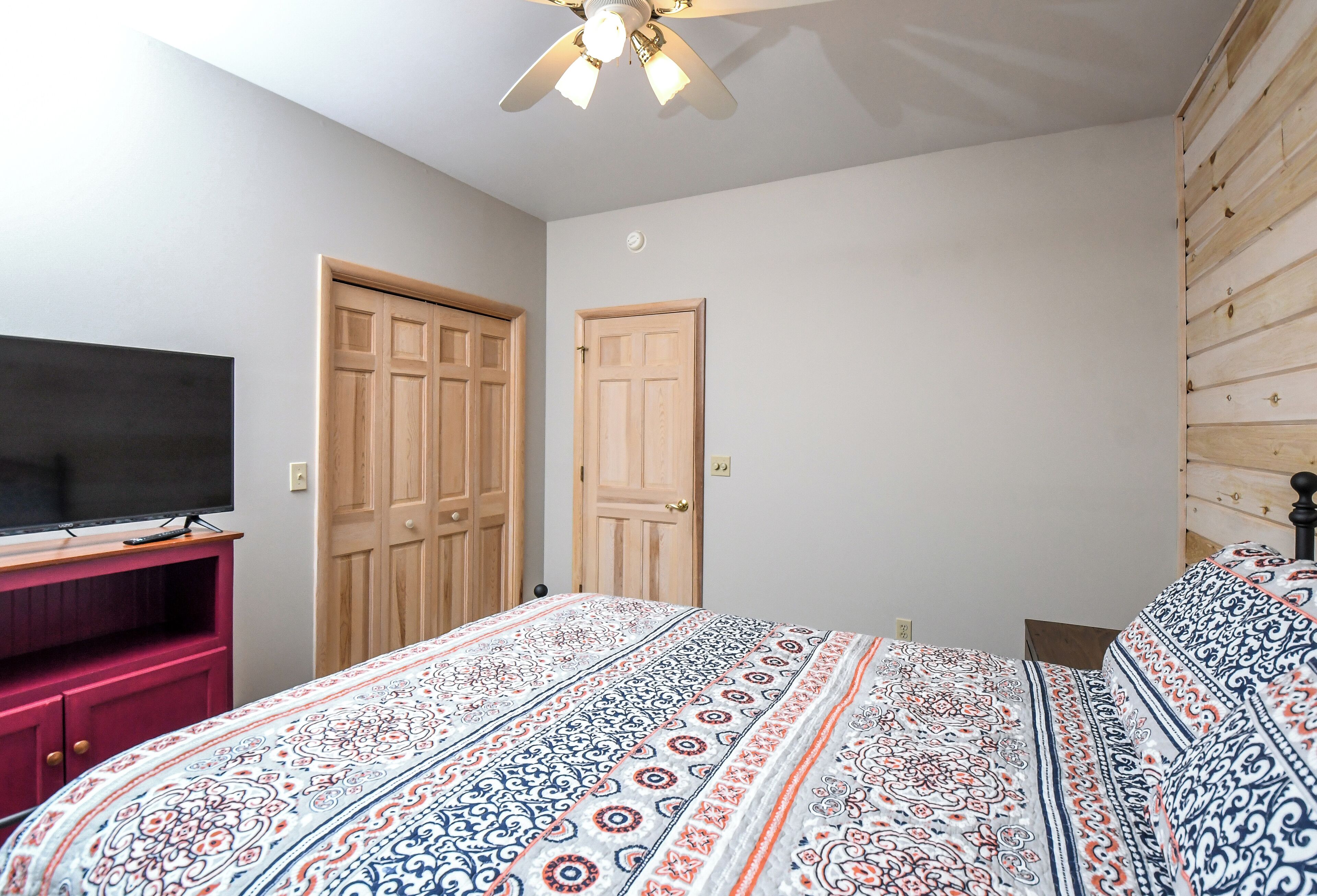 A bedroom with a bed, a TV on a stand, two wooden doors, and a ceiling fan.