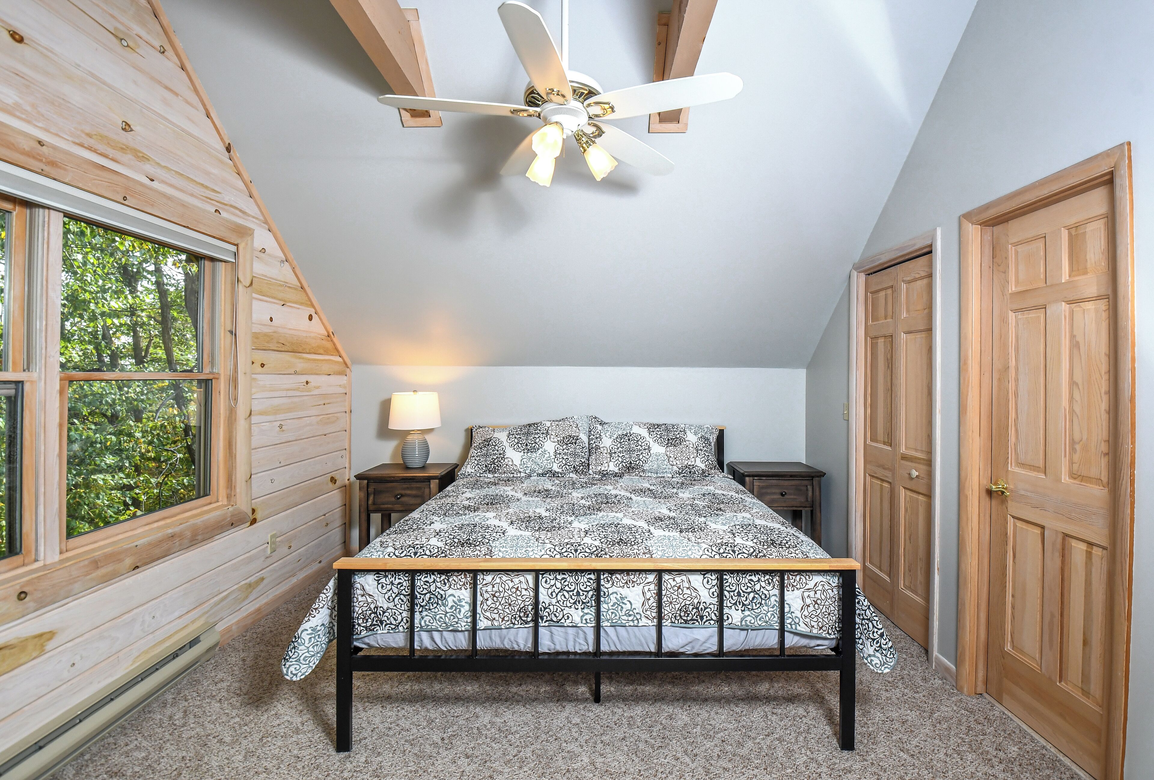 A bedroom with a sloped ceiling, a bed with patterned bedding, a ceiling fan, a nightstand with a lamp, and wooden doors.