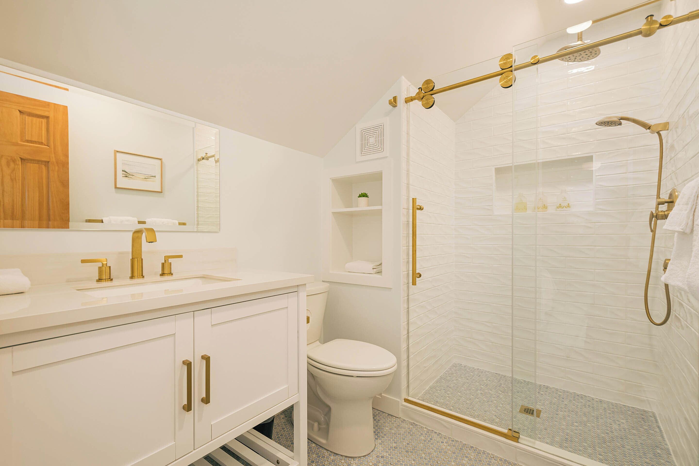 Un baño moderno con lavamanos blanco, accesorios dorados, un inodoro y zona de ducha con mampara de vidrio.