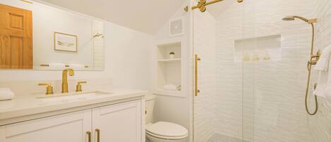 Un baño moderno con lavamanos blanco, accesorios dorados, un inodoro y zona de ducha con mampara de vidrio.