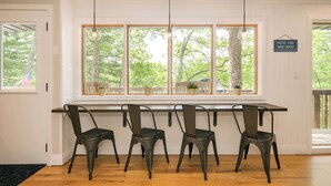 Uma sala de jantar com uma mesa comprida e quatro cadeiras, janelas amplas com vista para árvores, e um letreiro na parede.