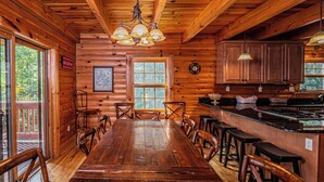 L’intérieur d’une cabane en bois comprend une grande table à manger, des chaises, un coin cuisine et une grande fenêtre.