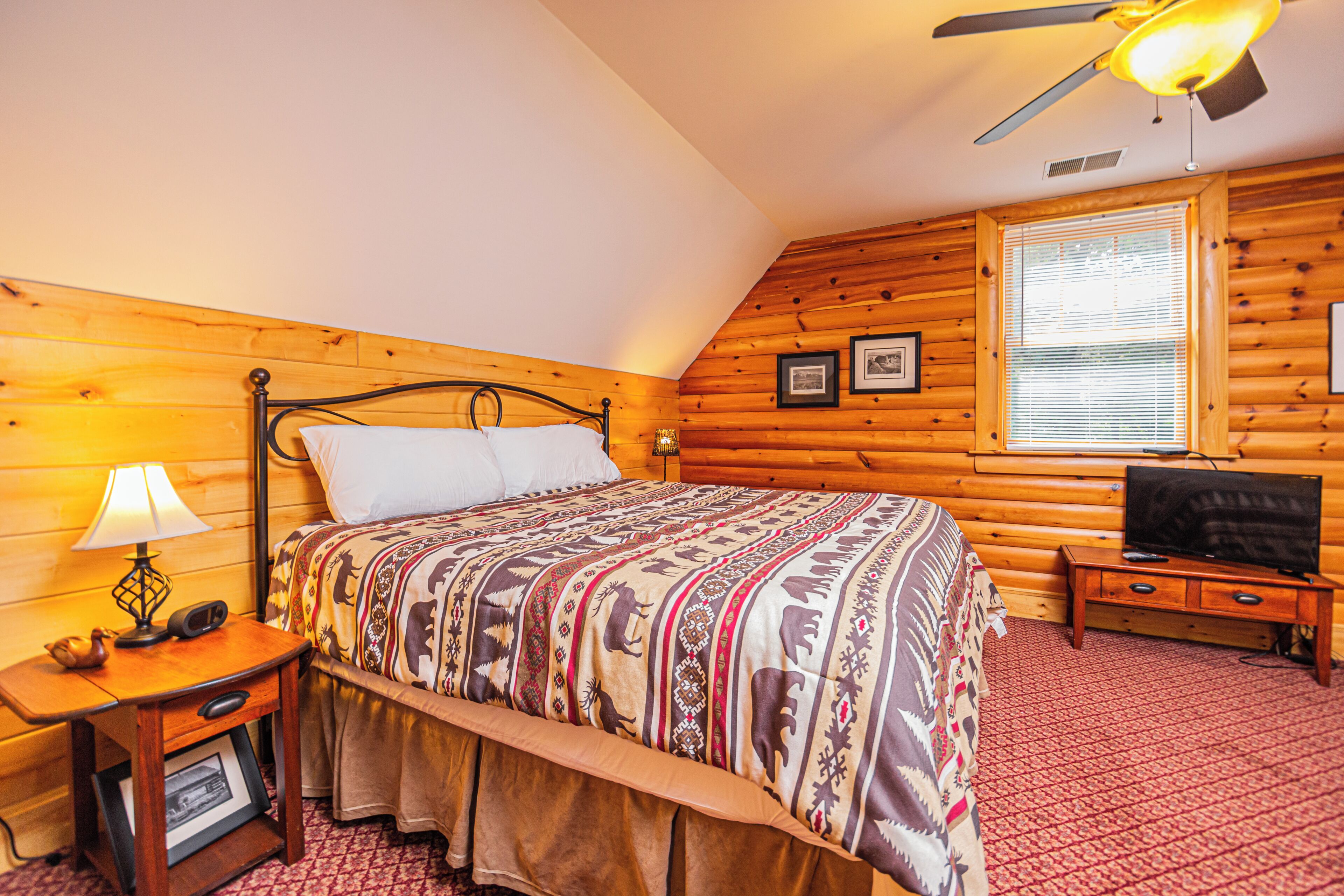 A cozy bedroom with a wooden log wall, a bed with patterned bedding, a nightstand with a lamp, a television, and a window with blinds.