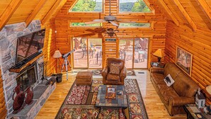 L’intérieur d’une cabane en rondins, avec une cheminée en pierre, des meubles en cuir et une grande fenêtre donnant sur une terrasse.