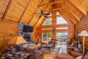 Interior de una cabaña de troncos con techo de madera, chimenea, mobiliario de cuero y un televisor.