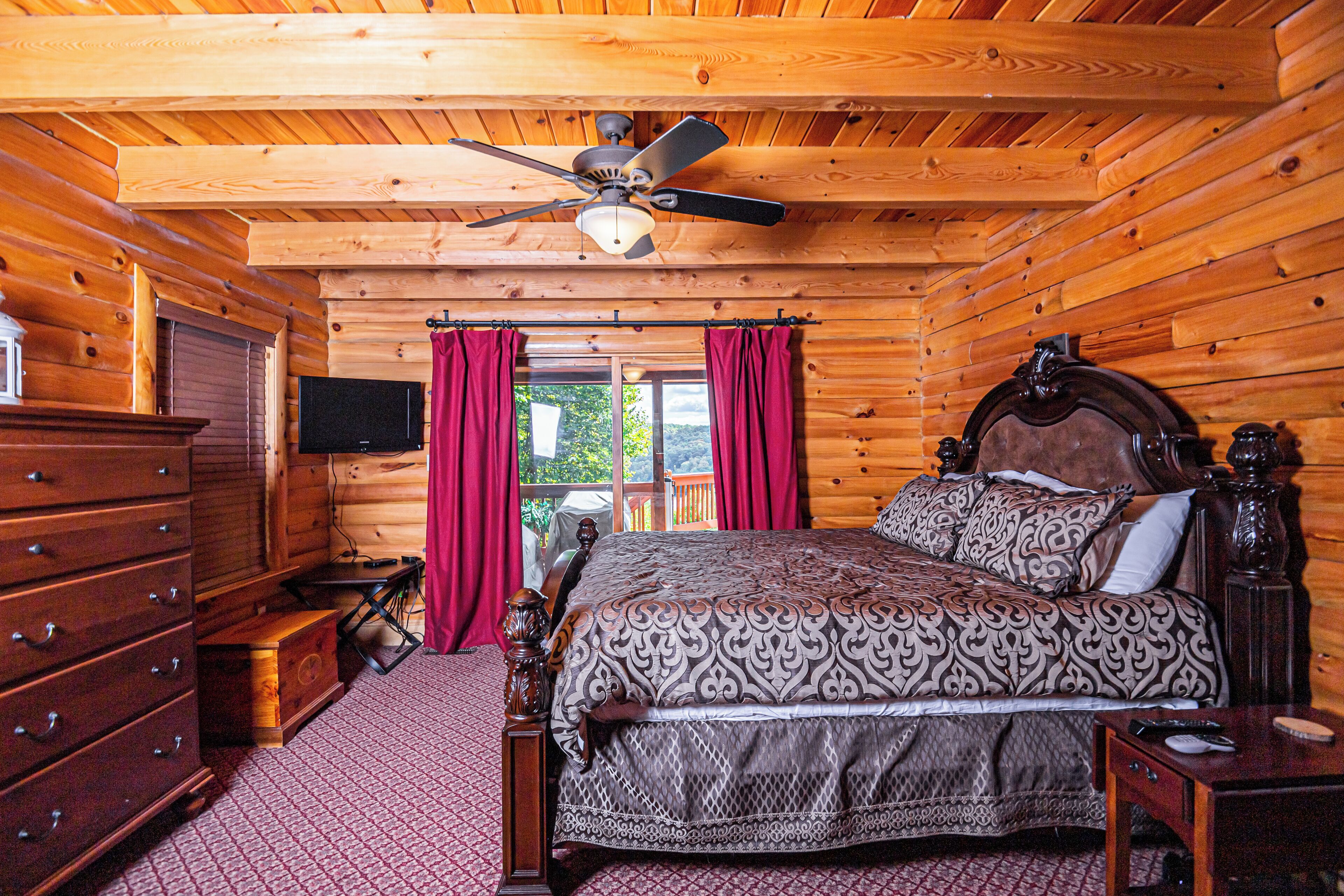 A log cabin bedroom with a wooden ceiling, a bed with patterned bedding, a dresser, and a television on the wall.