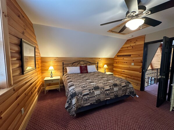 A bedroom with a wooden log wall, a bed with patterned bedding, two bedside lamps, a ceiling fan, and a red carpet. - Altitude Adjustment by Taylor Made Deep Creek (McHenry)