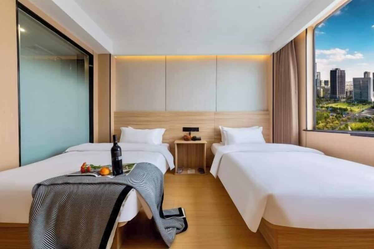A hotel room with two beds, a small table with a bottle and fruit, a nightstand, and a city view.