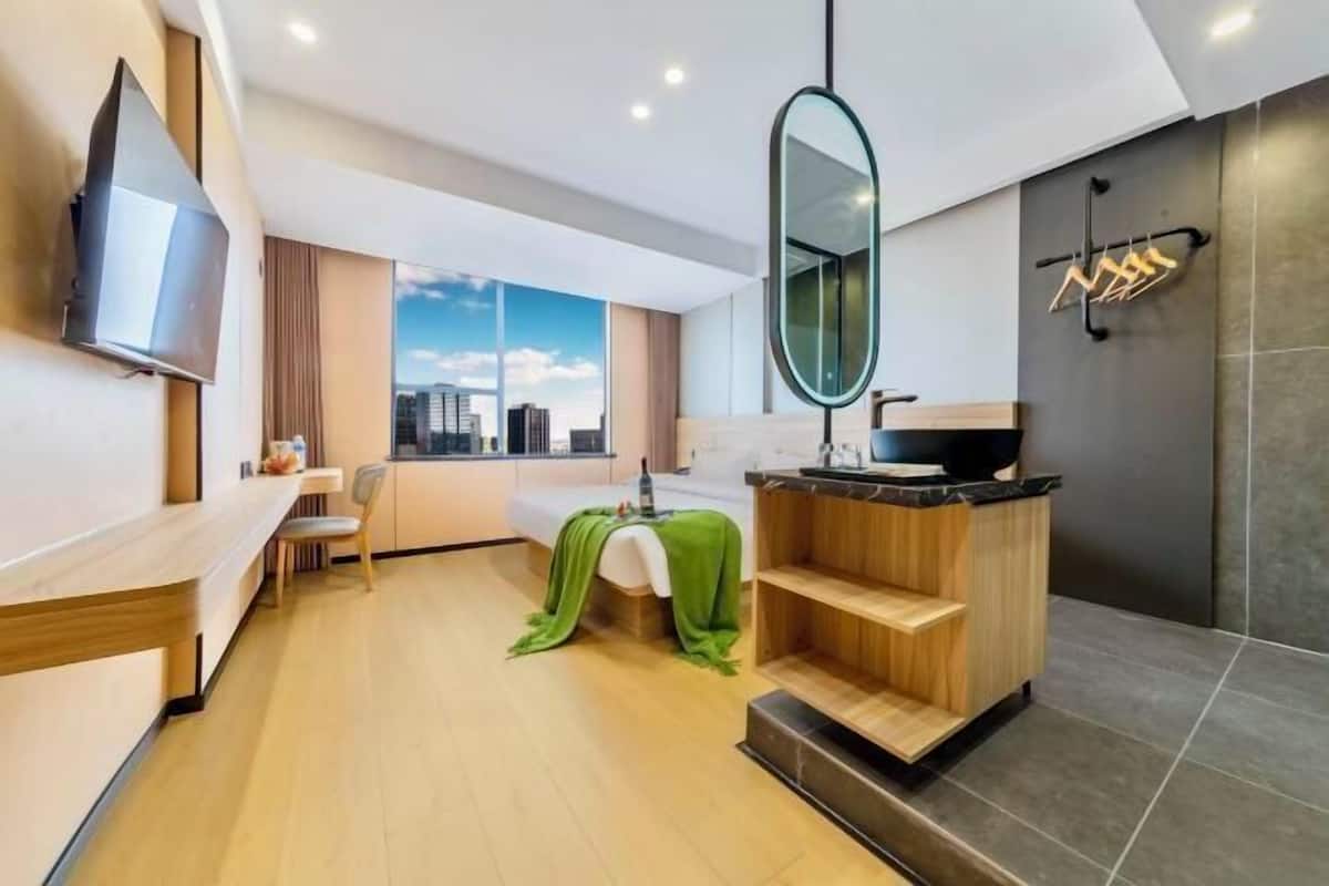 A modern hotel room with a wooden bench, a small kitchenette, a large mirror, and a view of the city.
