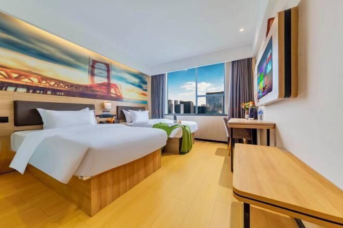 A hotel room with a bed, a desk, a chair, a window with a city view, and a painting on the wall.