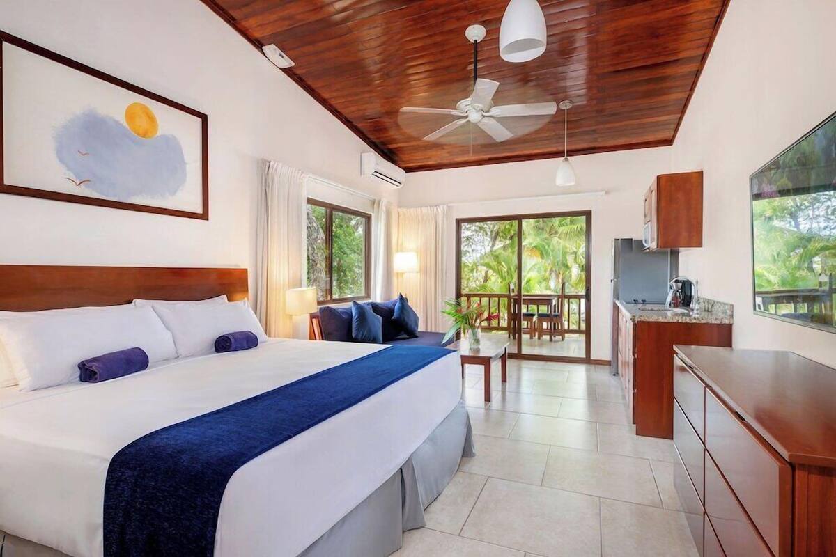 A hotel room with a large bed, a ceiling fan, a kitchenette, and a balcony with a view of greenery.