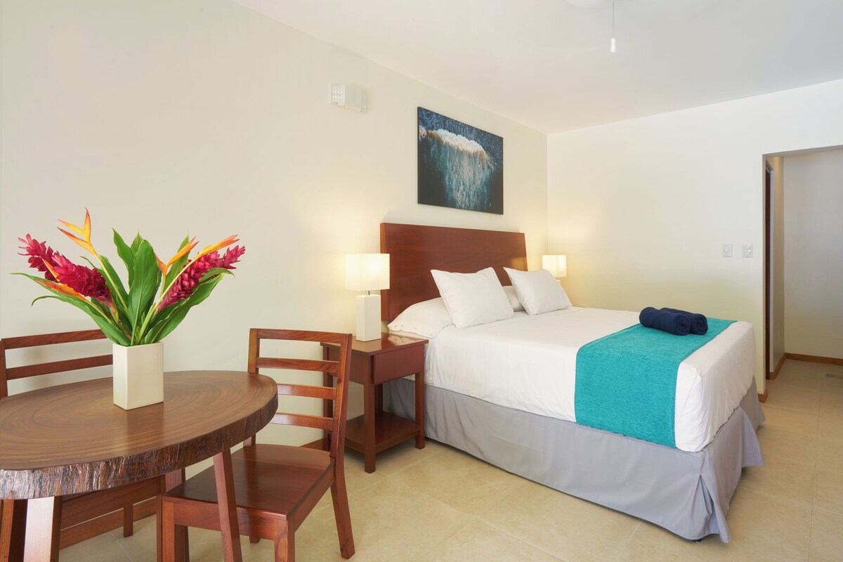Ein Hotelzimmer mit einem Bett, Nachttischen, einem Sessel, einem kleinen Tisch mit Blumenvase und einem Bild an der Wand.