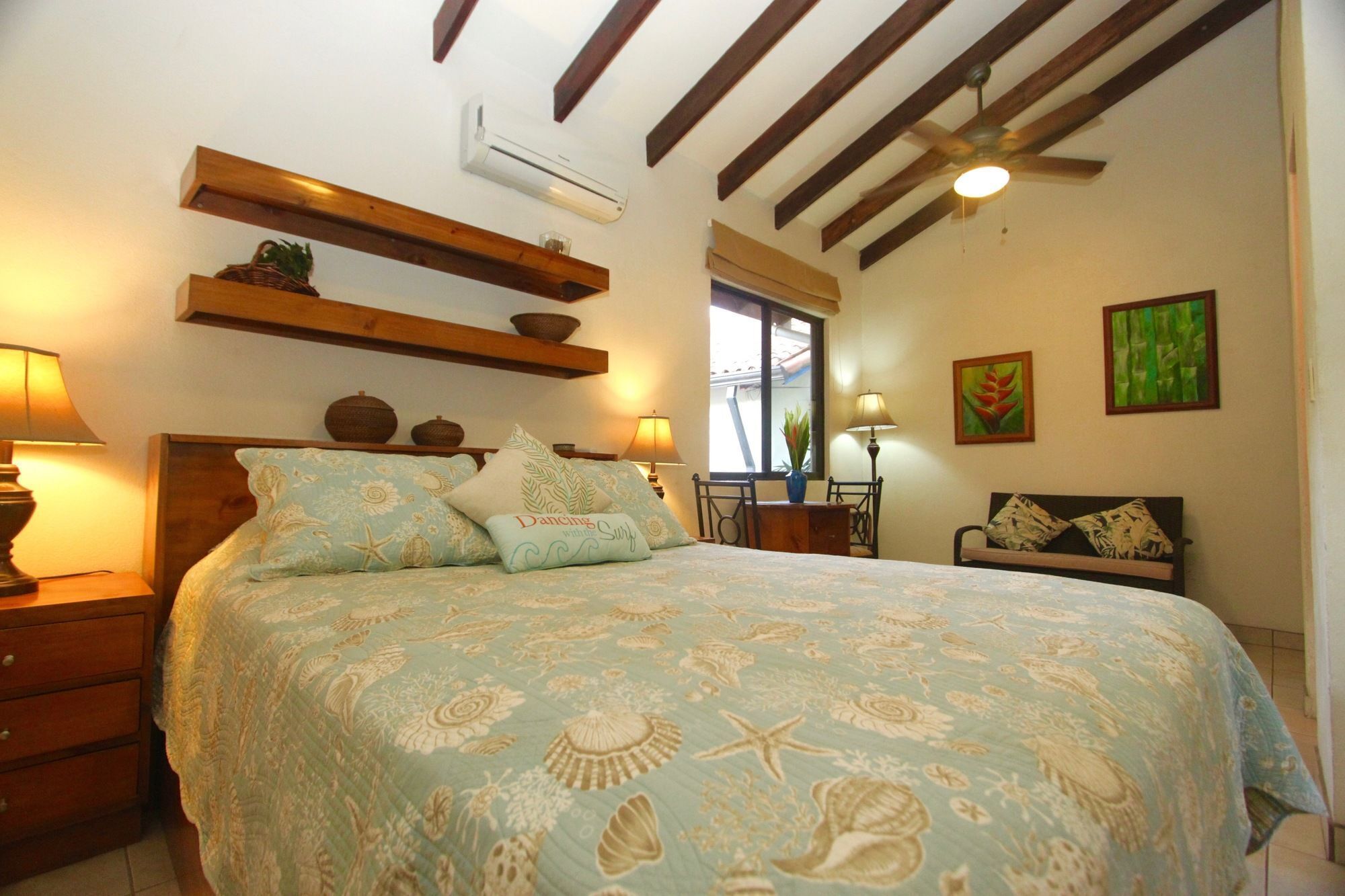 A bedroom with a bed, wooden beams, an air conditioner, a window, and two paintings on the wall.