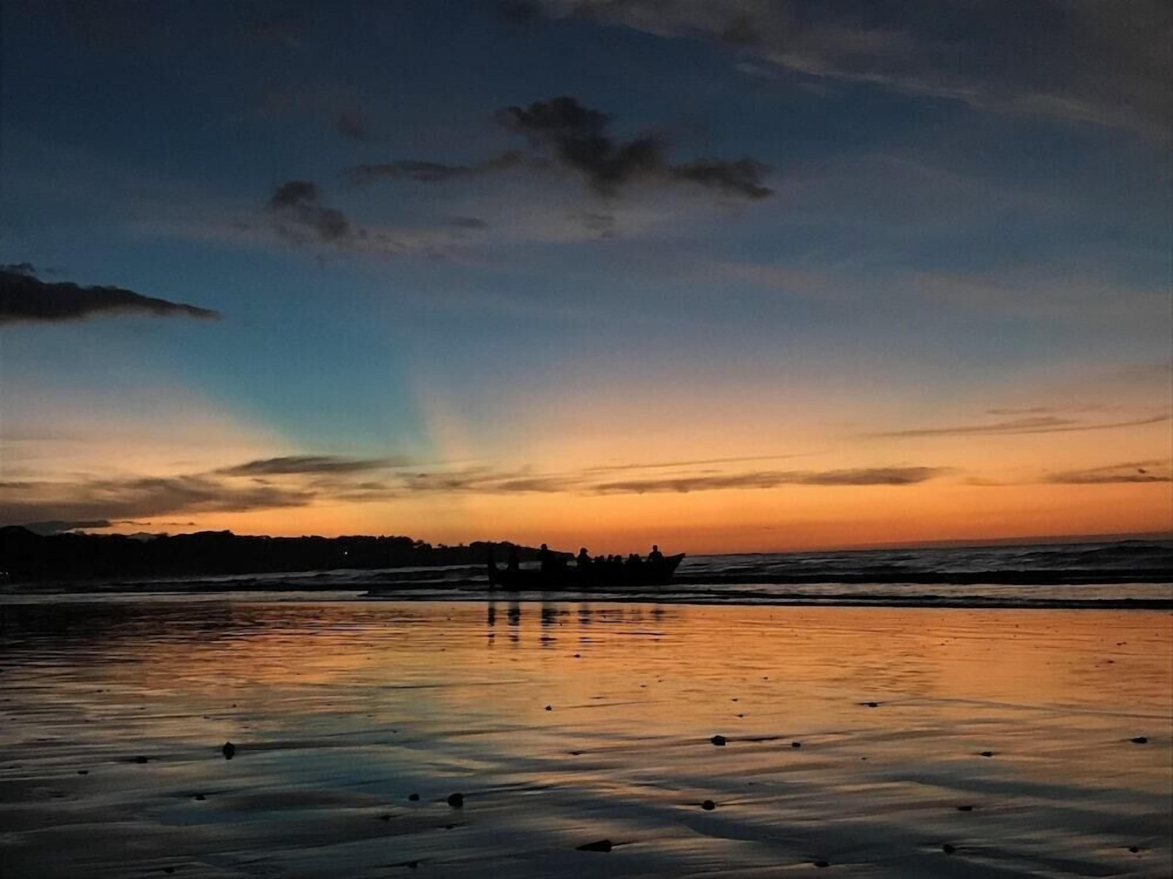 Une plage au coucher du soleil, avec un bateau et des personnes dont les silhouettes se détachent sur le ciel.