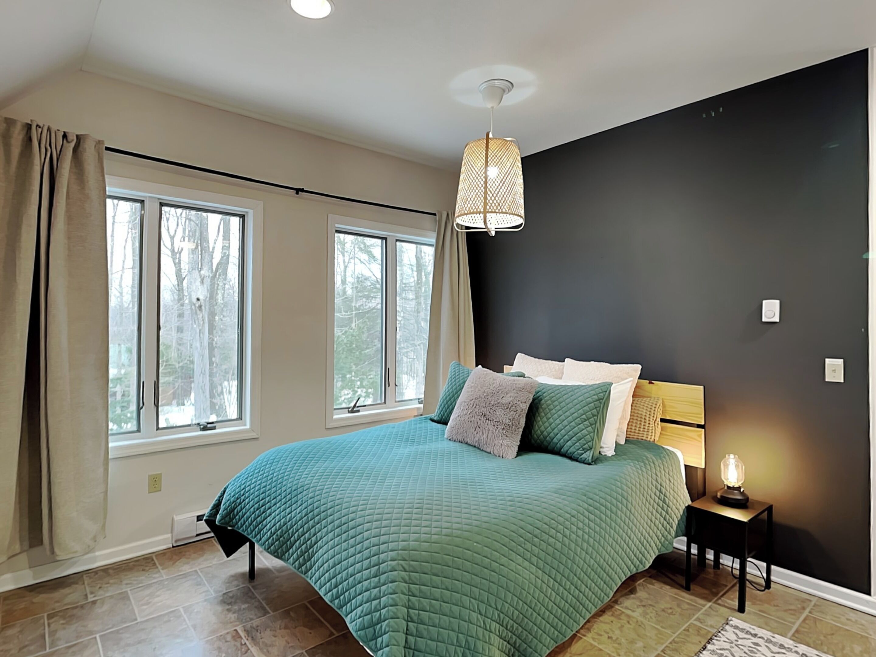A bedroom with a large bed, two windows, a black accent wall, and a bedside lamp.