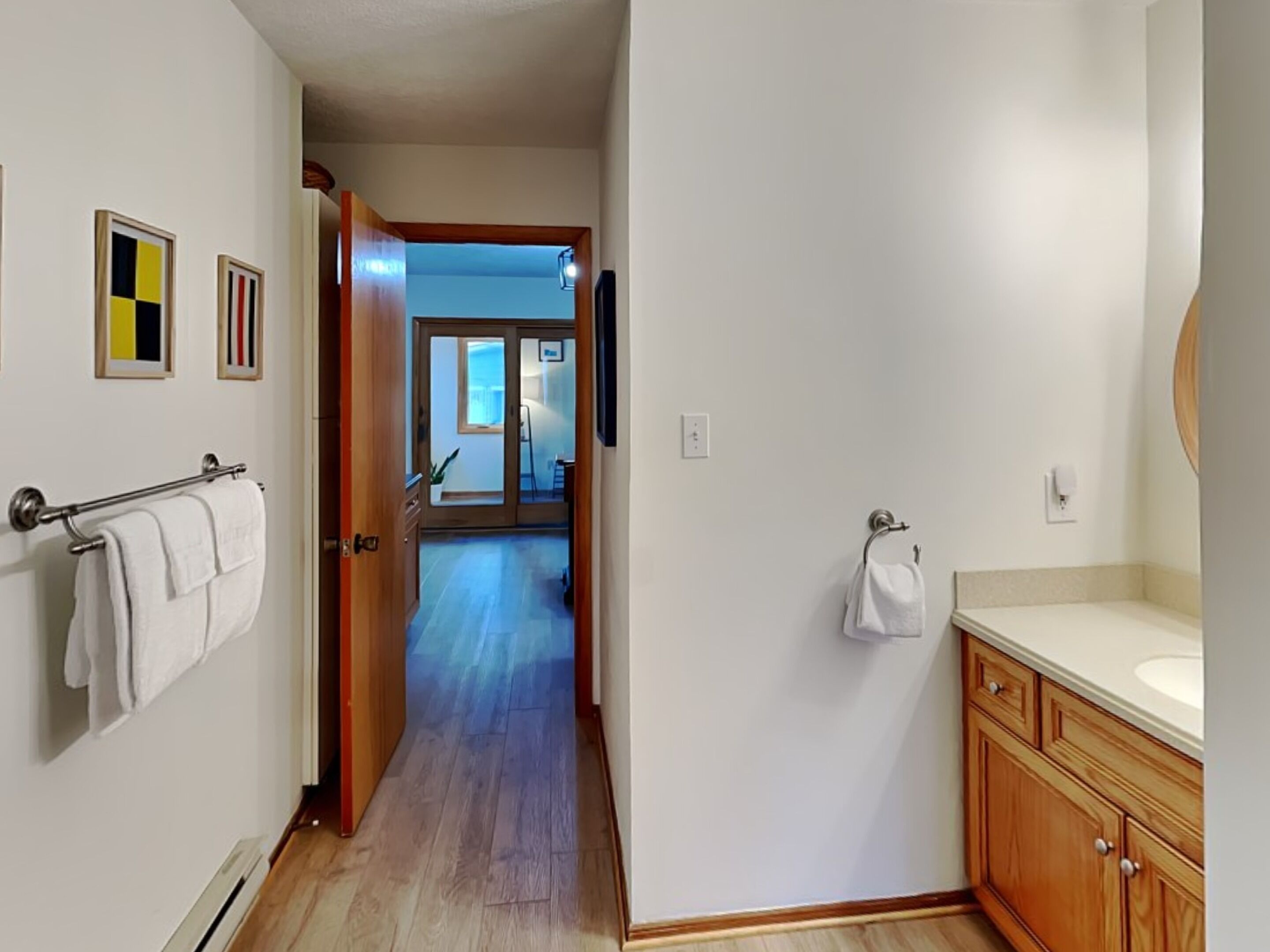 A hallway leading to a bathroom with a wooden vanity and a large mirror. There are framed pictures on the wall and a door leading to another room.