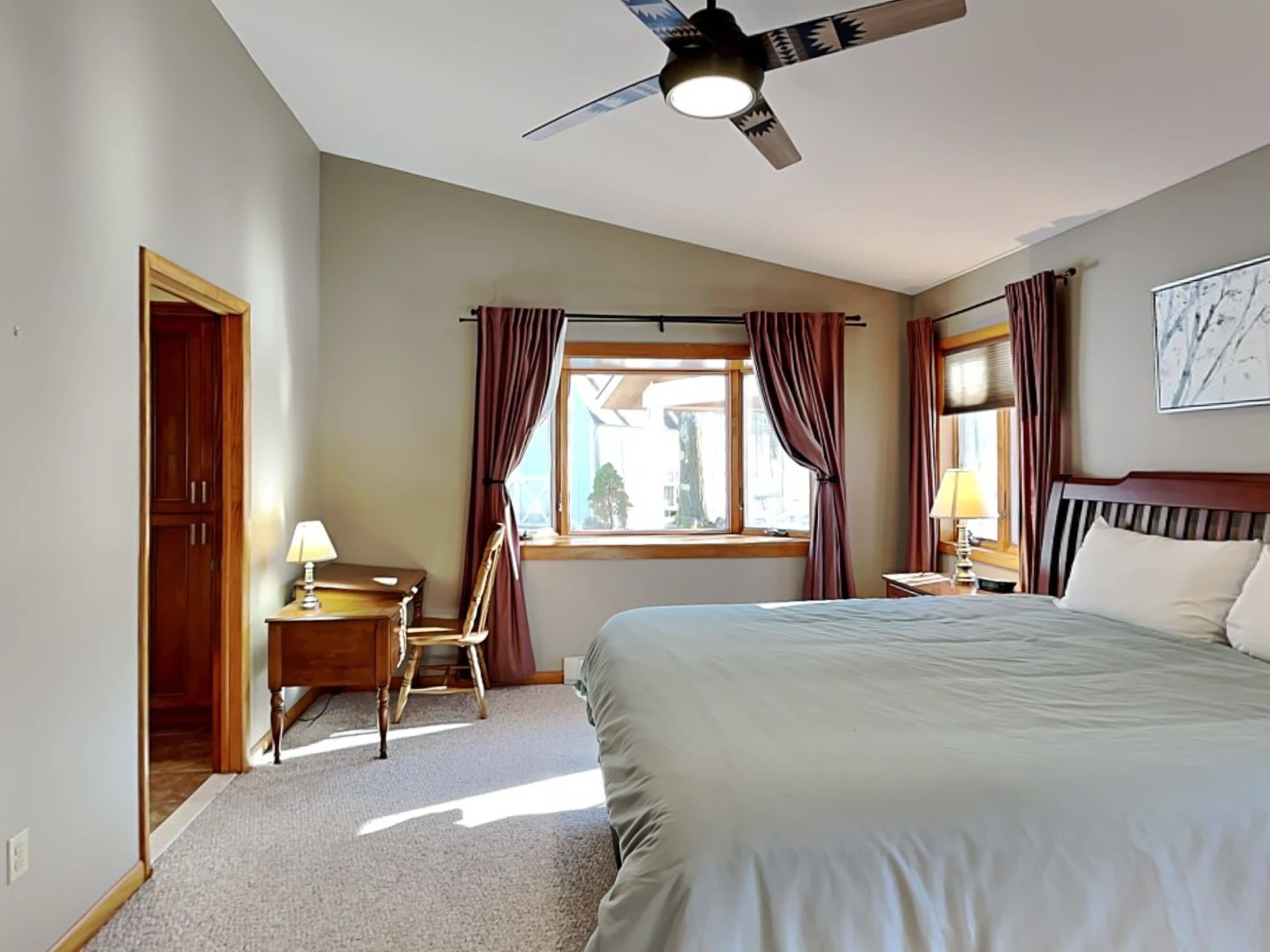 A hotel room with a bed, a desk with a lamp, a chair, a window with curtains, a ceiling fan, and a painting on the wall.