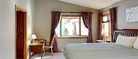 A hotel room with a bed, a desk with a lamp, a chair, a window with curtains, a ceiling fan, and a painting on the wall.