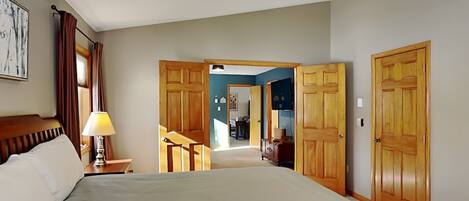 Une chambre d’hôtel avec un grand lit, un ventilateur de plafond, des portes en bois et un couloir menant à une autre chambre.
