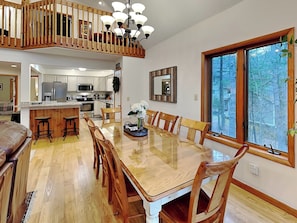A spacious dining area with a wooden table and chairs, a kitchen in the background, and a large window with a view of trees. - American Pride by Taylor Made Deep Creek (Oakland)