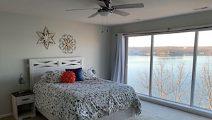 Quarto com uma janela grande, ventoinha no teto e cama com roupa de cama estampada.