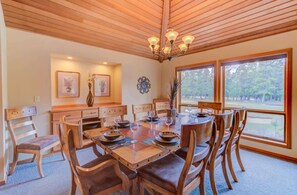 A dining room with a wooden table set for four, surrounded by chairs. There is a chandelier above the table, and the room has large windows offering a view of the outdoors. - Amazing view of golf course, hot tub, AC, pets ok (Sunriver)