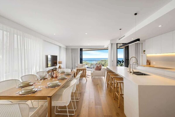 A modern kitchen with a large island, dining area with a view, and a living space with a sofa and a TV. - Coolum First Bay 121 131 (Coolum Beach)