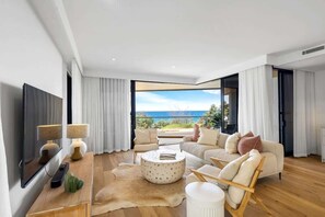 A modern living room with a large TV, a round coffee table, a sofa, and a view of the sea.