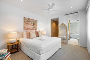 A modern bedroom with a large bed, bedside lamps, a ceiling fan, a decorative wall piece, and a bathroom with a bathtub and mirror.