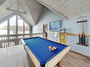 A pool table in a room with wooden beams on the ceiling, a fan, and a window with a view of trees. - At Lake's Edge by Taylor Made Deep Creek (McHenry)