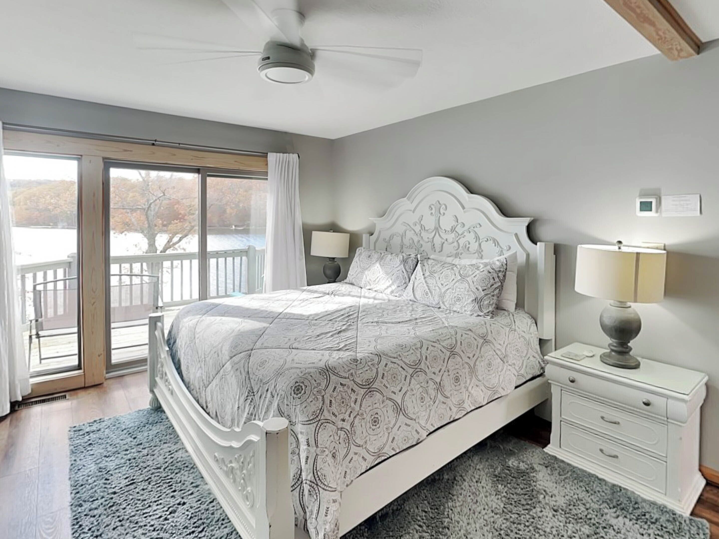 A bedroom with a large bed, a nightstand, a ceiling fan, and a view of a balcony.