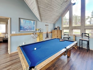 A pool table in a room with wooden floors, a ceiling fan, and a window with a view of trees. - At Lake's Edge by Taylor Made Deep Creek (McHenry)