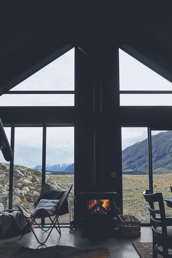 A cozy room with a fireplace, large windows, and a view of mountains. - Secluded Rustic Cabin near Twizel (Ben Ohau)