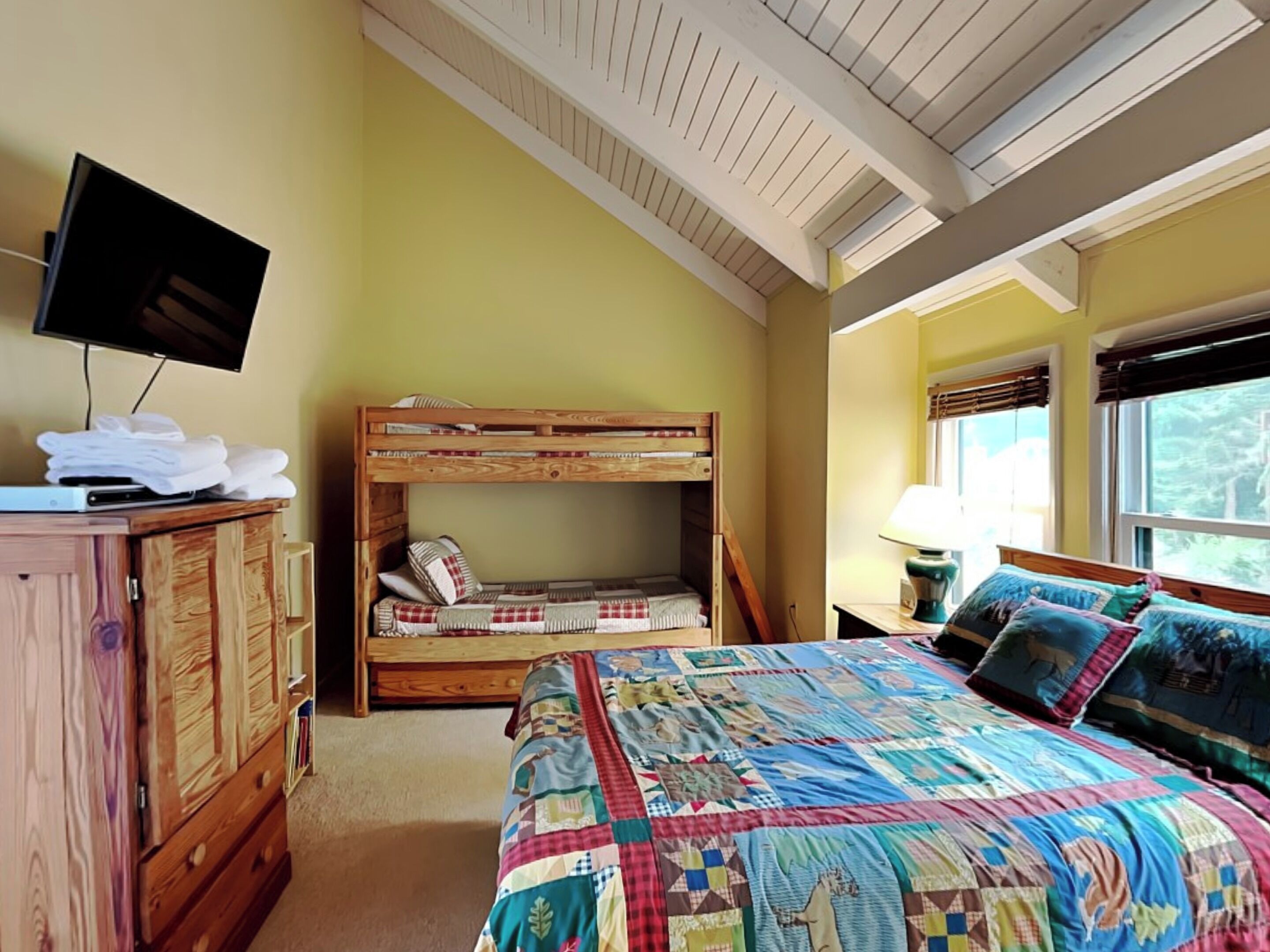 Une chambre avec un lit superposé, un téléviseur posé sur un meuble en bois et une fenêtre avec des stores.