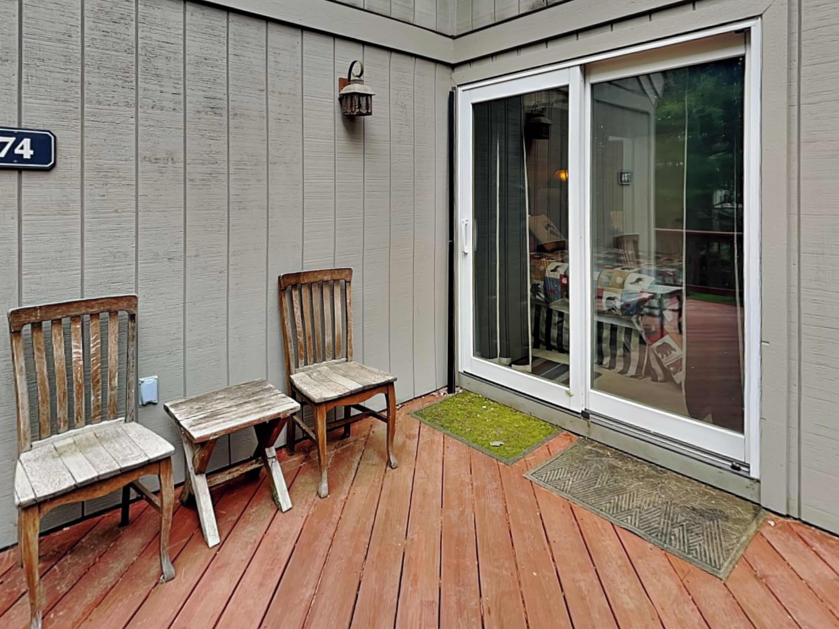 A wooden deck with two chairs and a small table, a sliding glass door, and a number '74' sign on the wall.