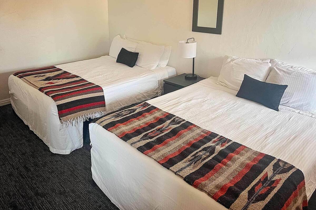 Two single beds with white linens and striped blankets, a bedside table with a lamp, and a wall-mounted mirror.