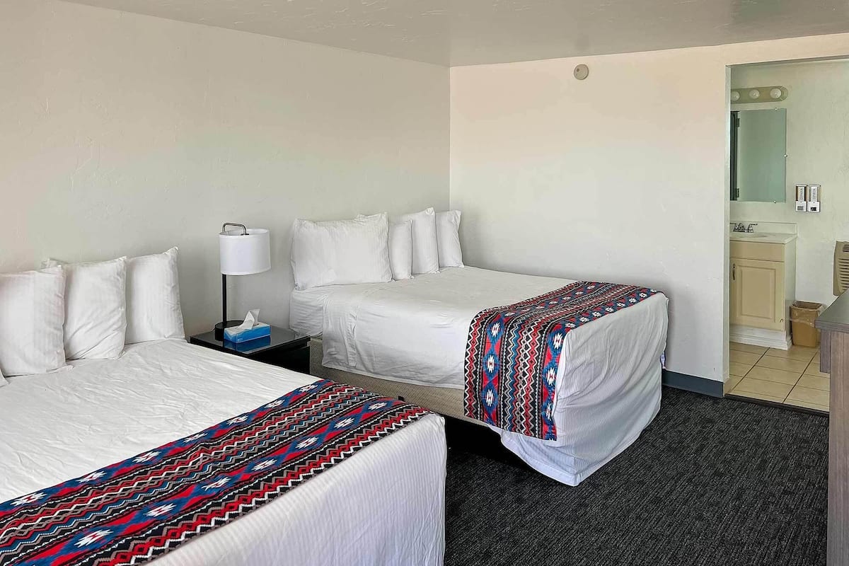 A hotel room with two beds, a nightstand with a lamp, a patterned throw blanket, and a bathroom visible through an open door.
