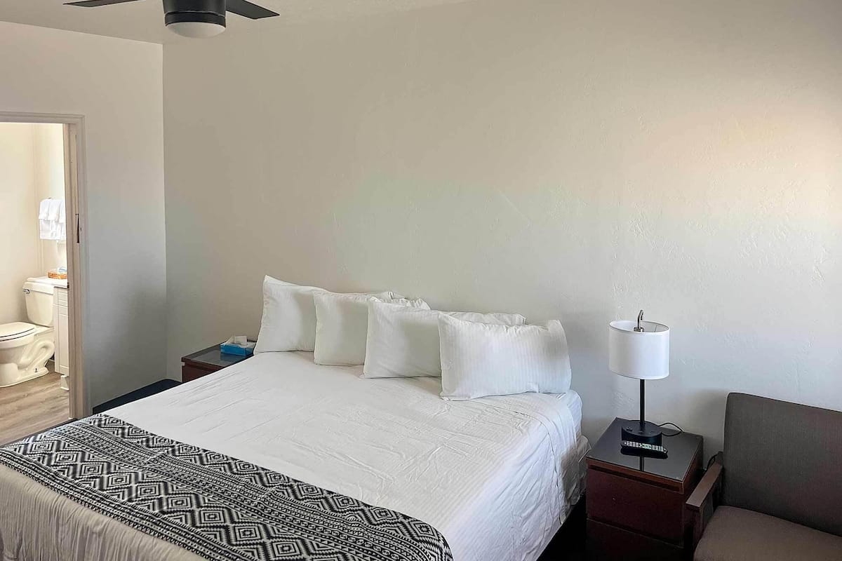 A hotel room with a bed, a bedside table with a lamp, a chair, a ceiling fan, and a view of the bathroom.