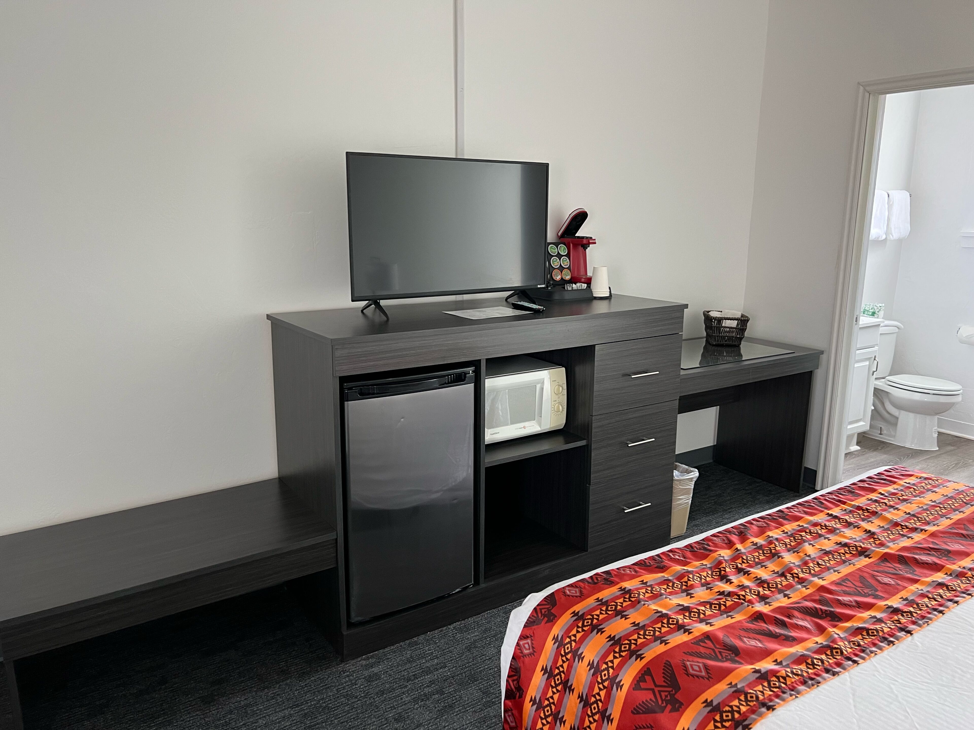 A hotel room with a dark wooden desk, a flat-screen TV, a microwave, and a small refrigerator.