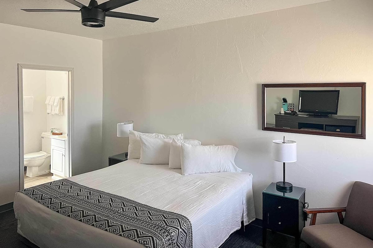A hotel room with a bed, a nightstand, a chair, a desk, a ceiling fan, a mirror, and a bathroom visible through an open door.