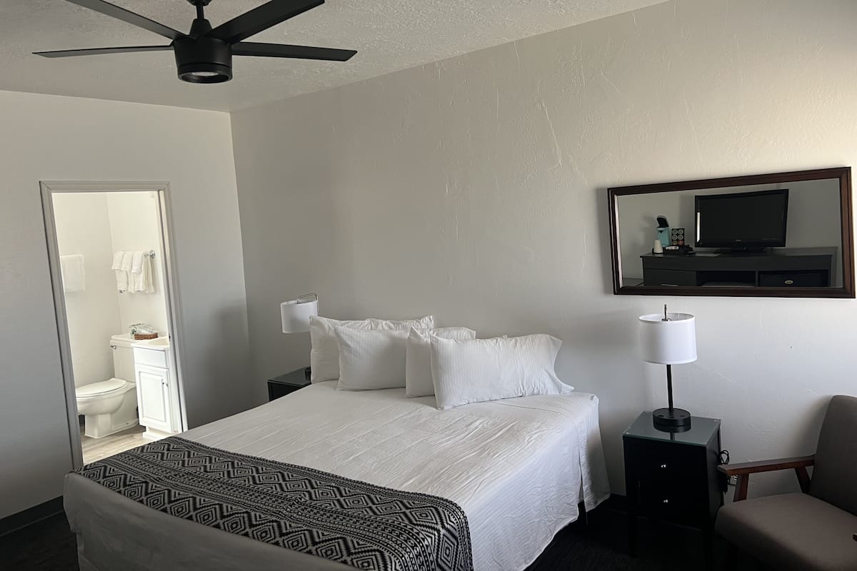 A hotel room with a bed, a ceiling fan, a bedside table with a lamp, a chair, a TV, and a bathroom visible through an open door.
