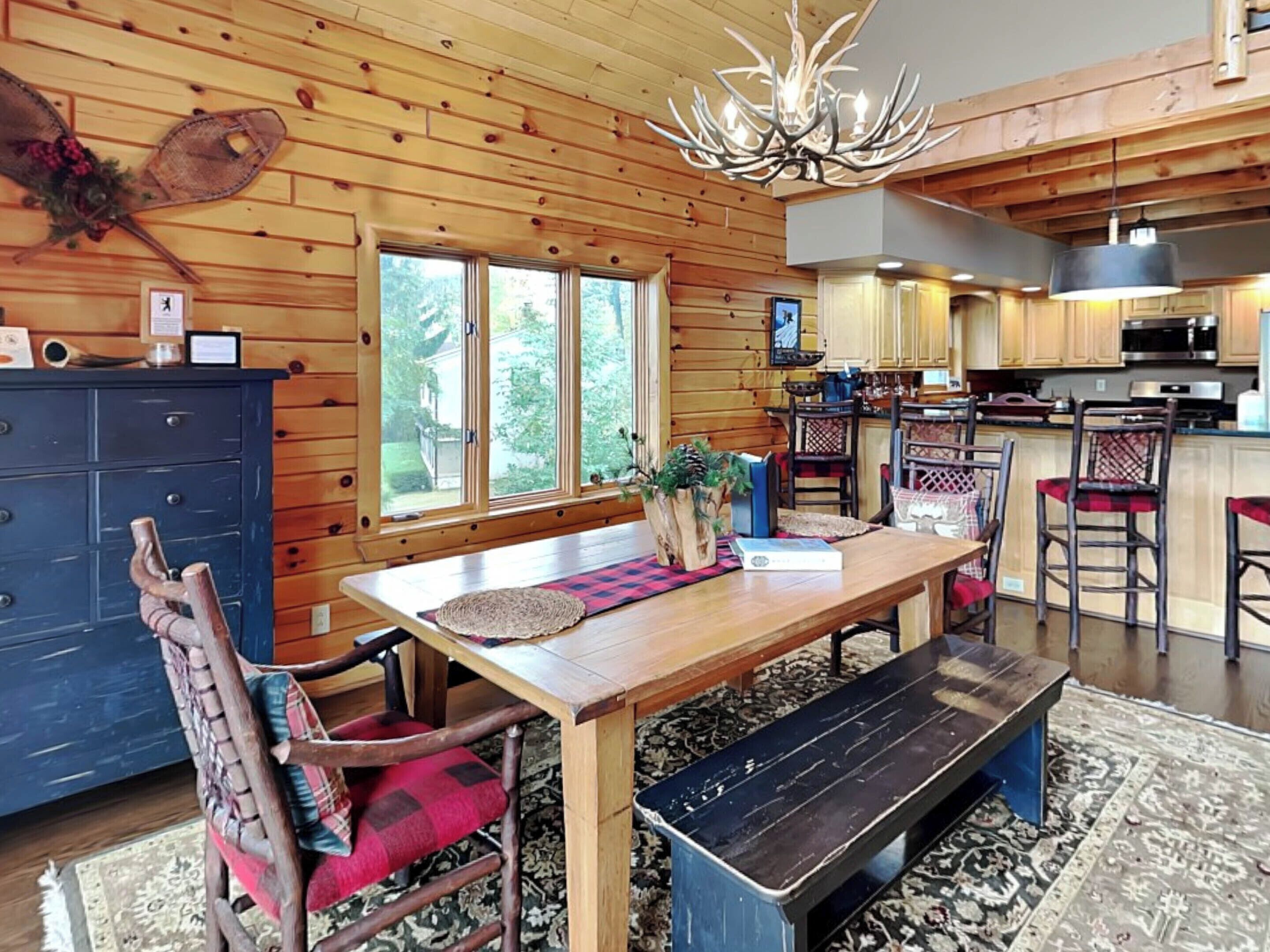 A wooden cabin interior with a dining area, kitchen, and large windows.