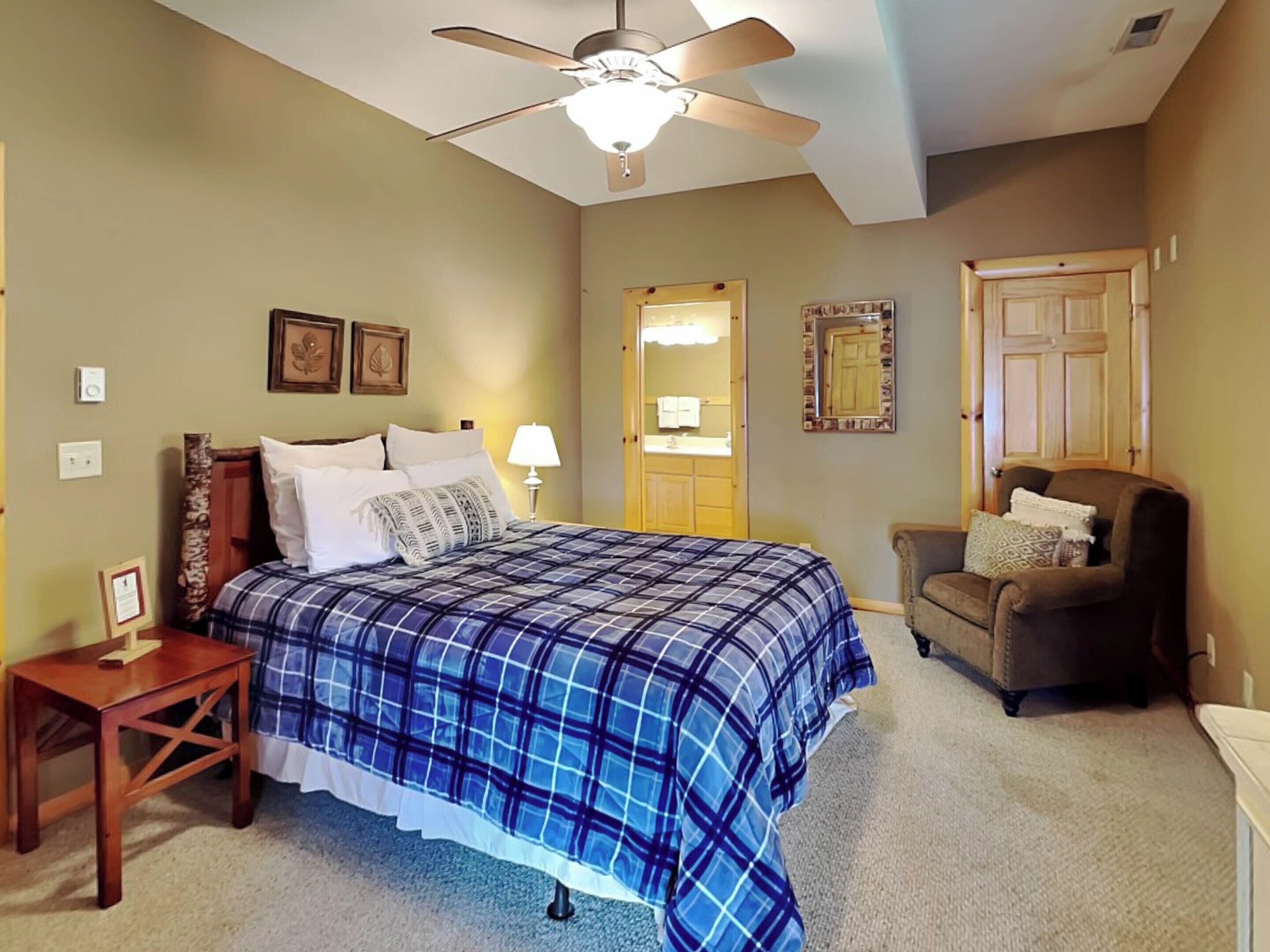 A bedroom with a bed, a chair, a nightstand, a ceiling fan, a mirror, and a door.