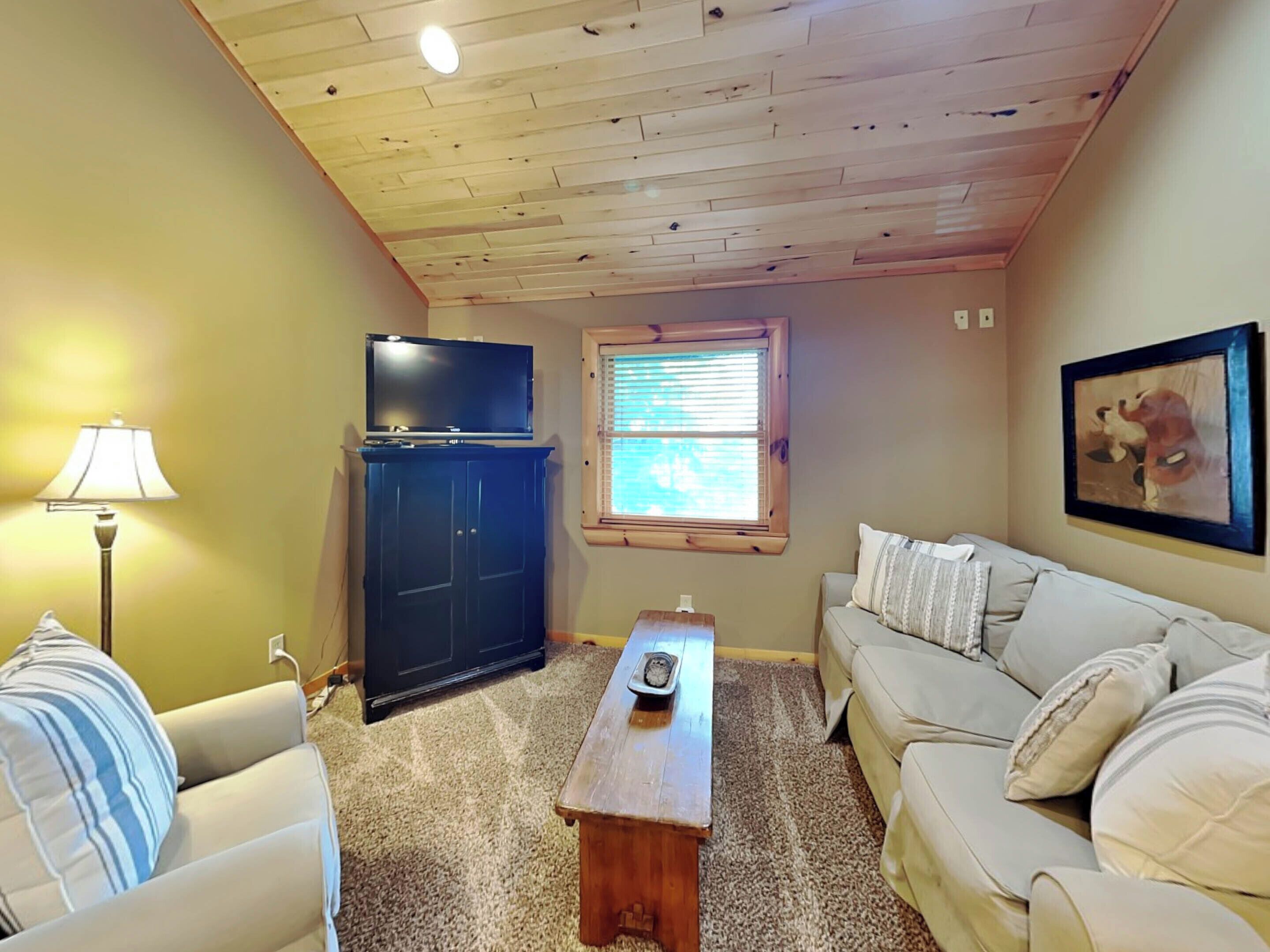 A living room with a wooden ceiling, a television on a cabinet, a window with blinds, a sofa, and a lamp.