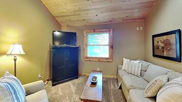 A living room with a wooden ceiling, a television on a cabinet, a window with blinds, a sofa, and a lamp.