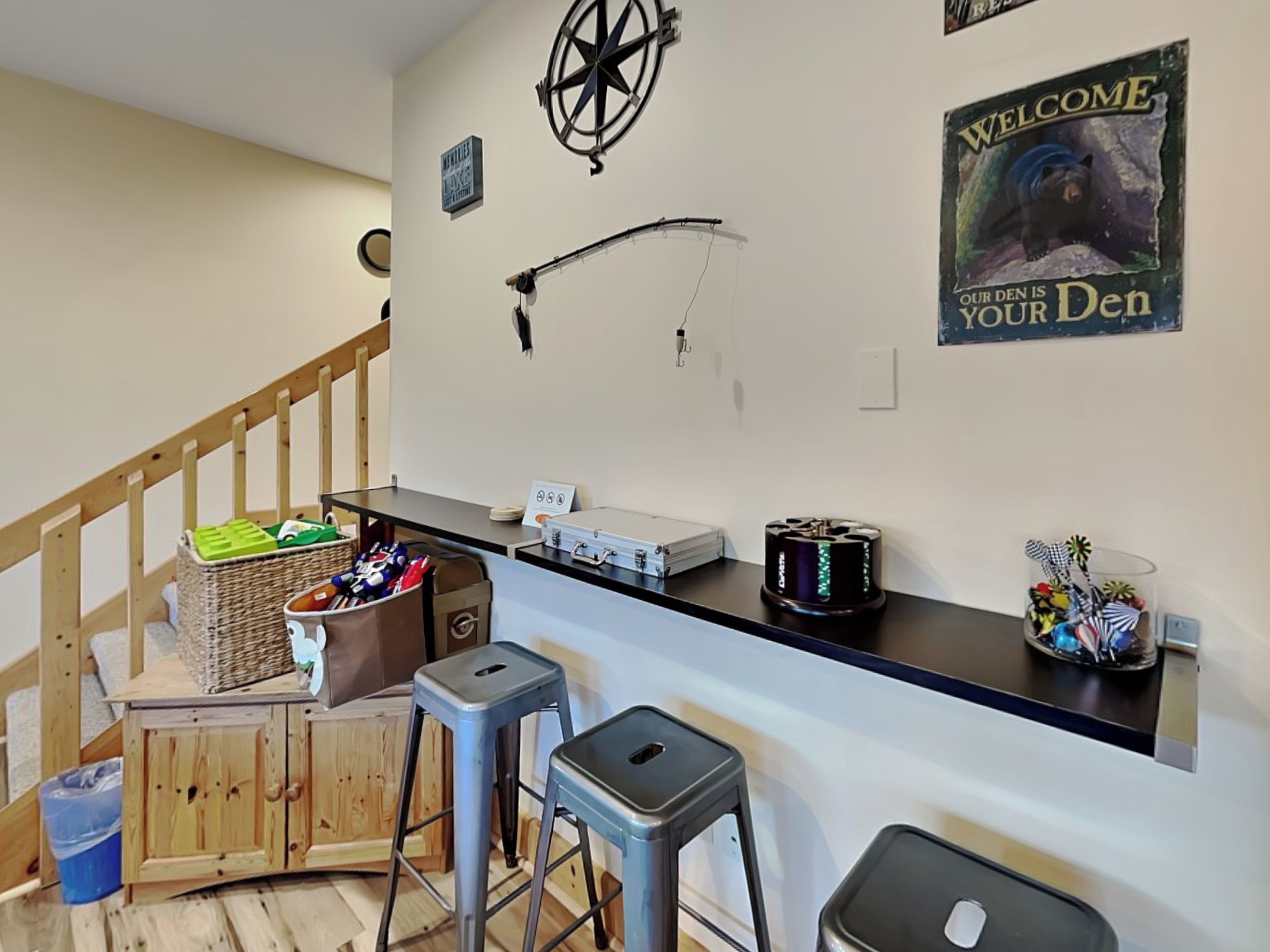 A room with a wooden staircase, a wall-mounted compass, a welcome sign, a desk with a computer, and a basket of items.