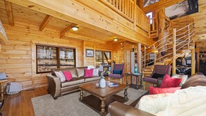L’intérieur d’un chalet en bois, avec un canapé, des fauteuils, une table basse et un escalier.